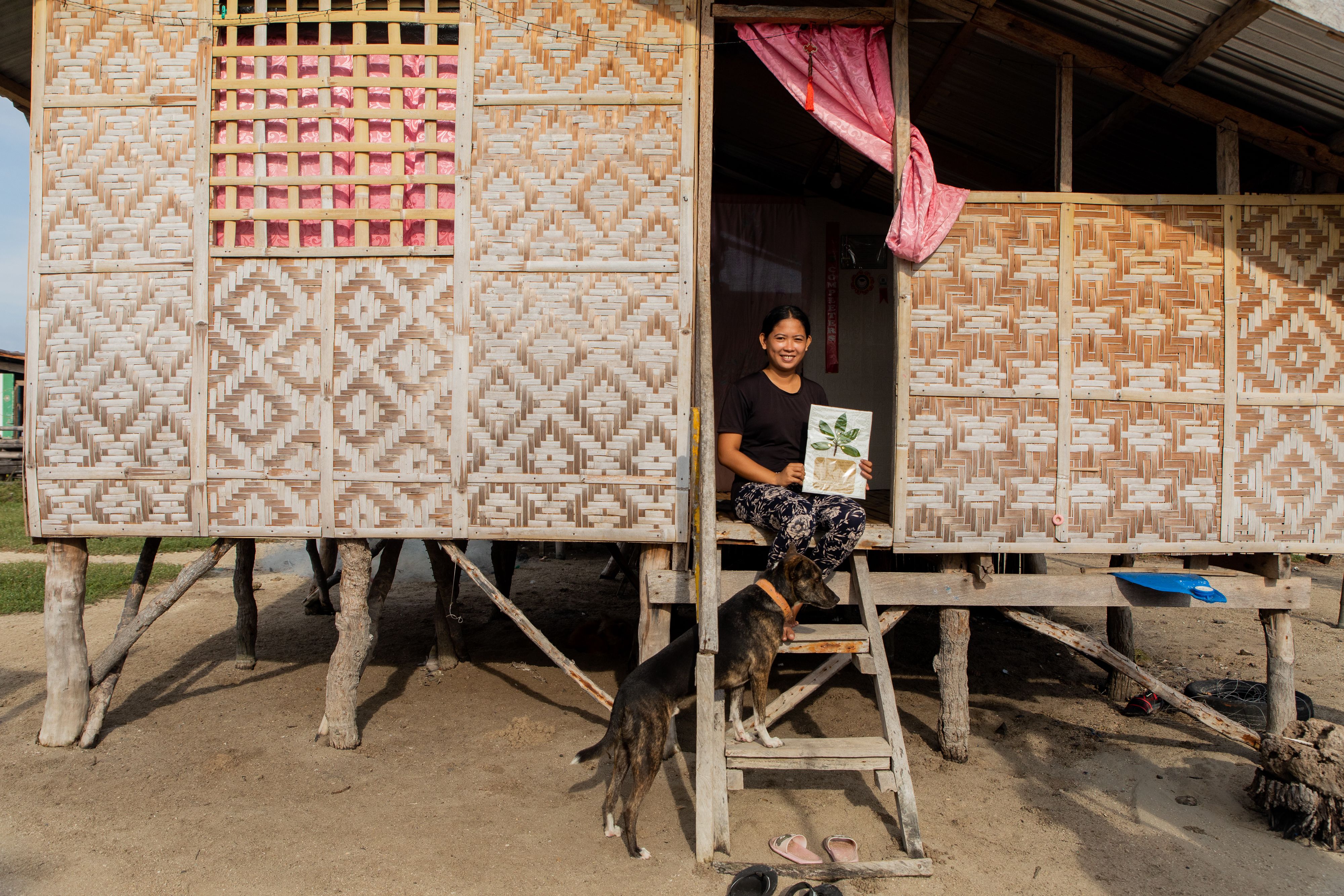 A portrait of Hazel Brum sitting in the doorway to her house, holding her object of memory: a bakhaw (mangrove) stem