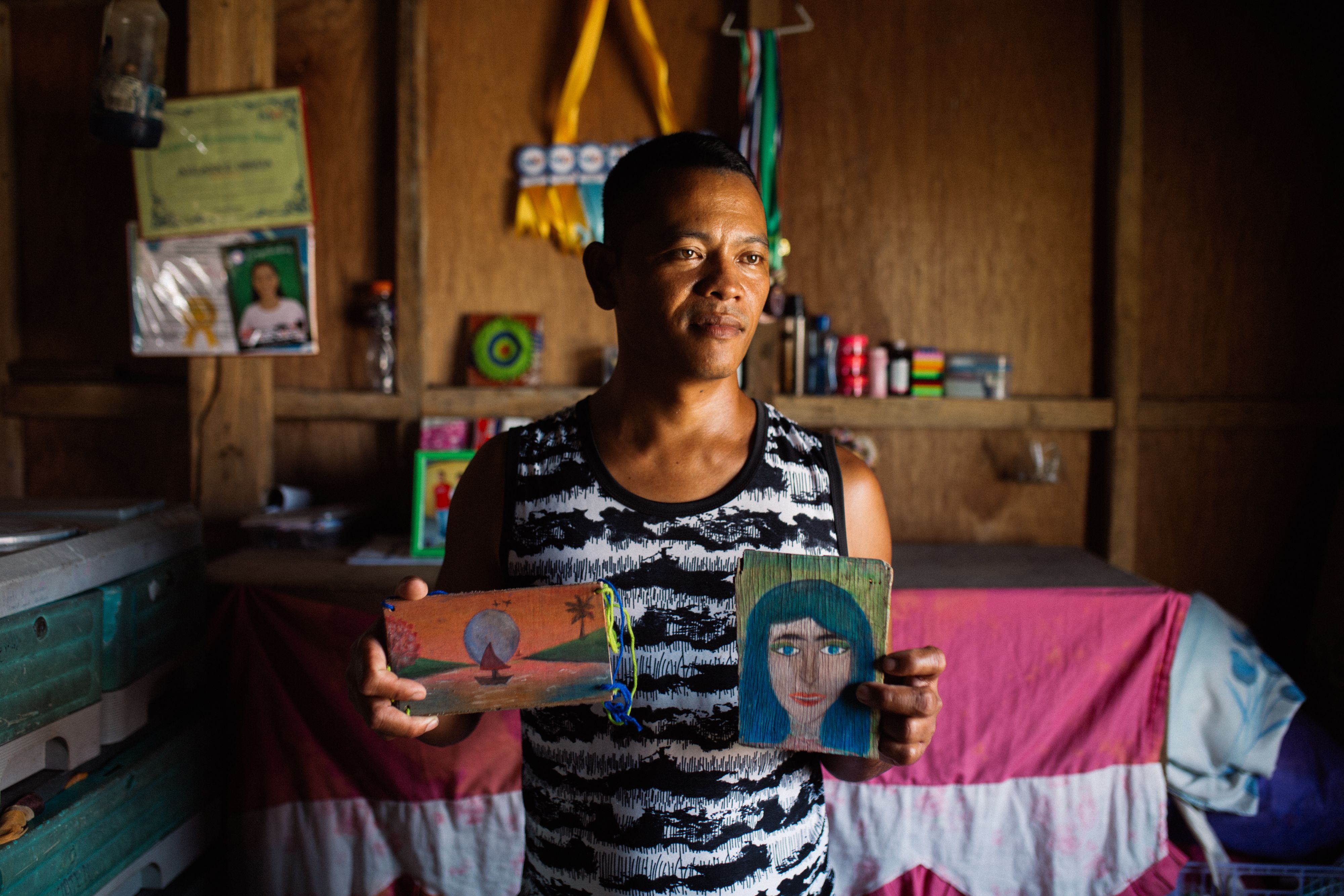 A photo of Arnold Obguia holding his objects of memory: paintings on broken pieces of wood